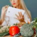 Blonde woman using tablet with fresh vegetables like broccoli and tomatoes in a kitchen setting.