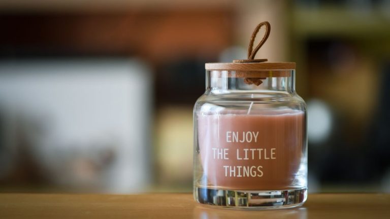 A glass jar candle with 'Enjoy the little things' text and wooden lid on table.